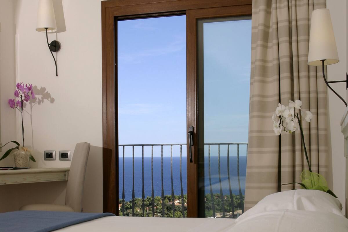Habitación de hotel con cama, escritorio con lámpara, teléfono y vista al mar desde el balcón.