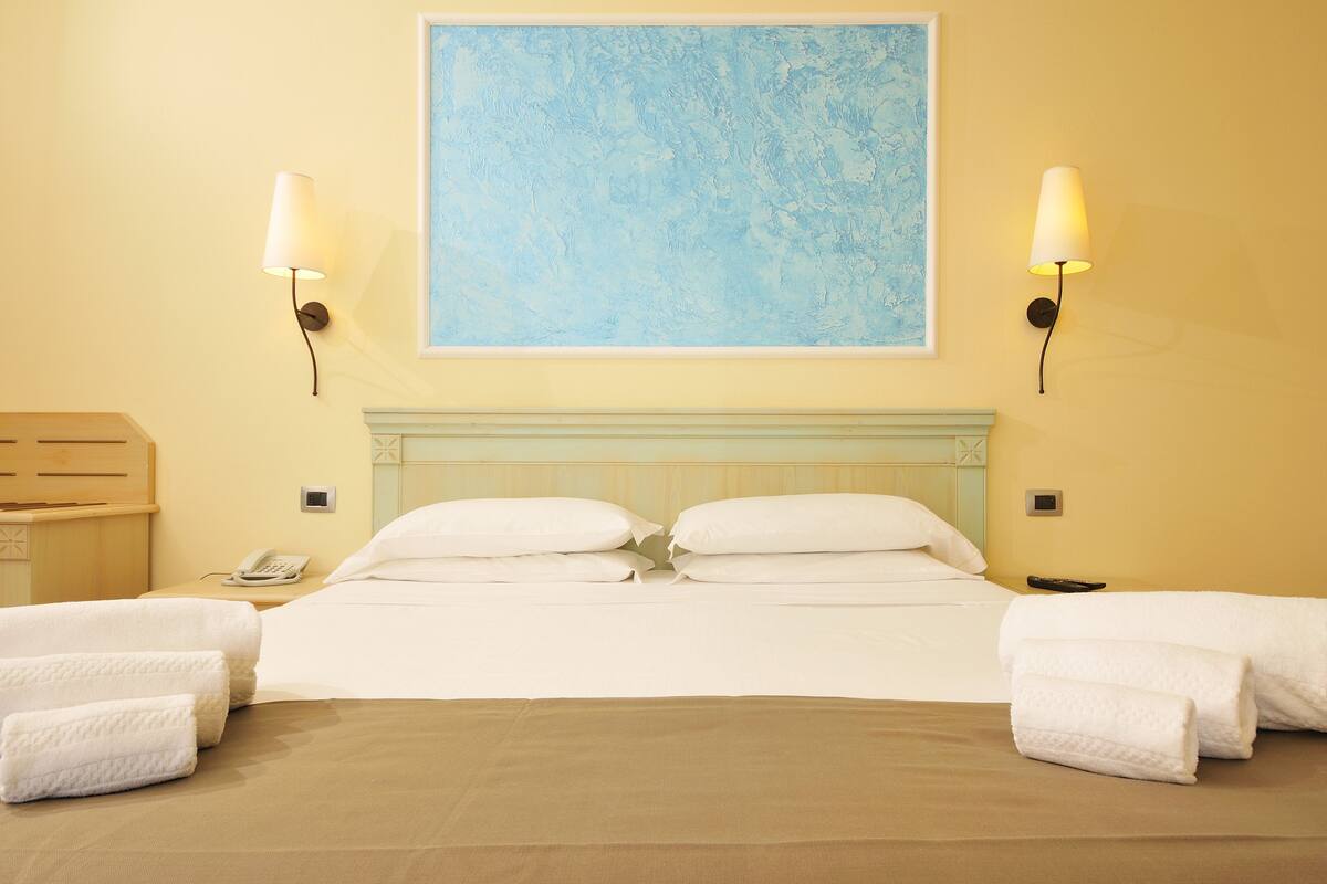 Una habitación de hotel con una cama grande, dos lámparas fijadas a la pared, un cuadro sobre la cama y una mesita de noche de madera con un teléfono.