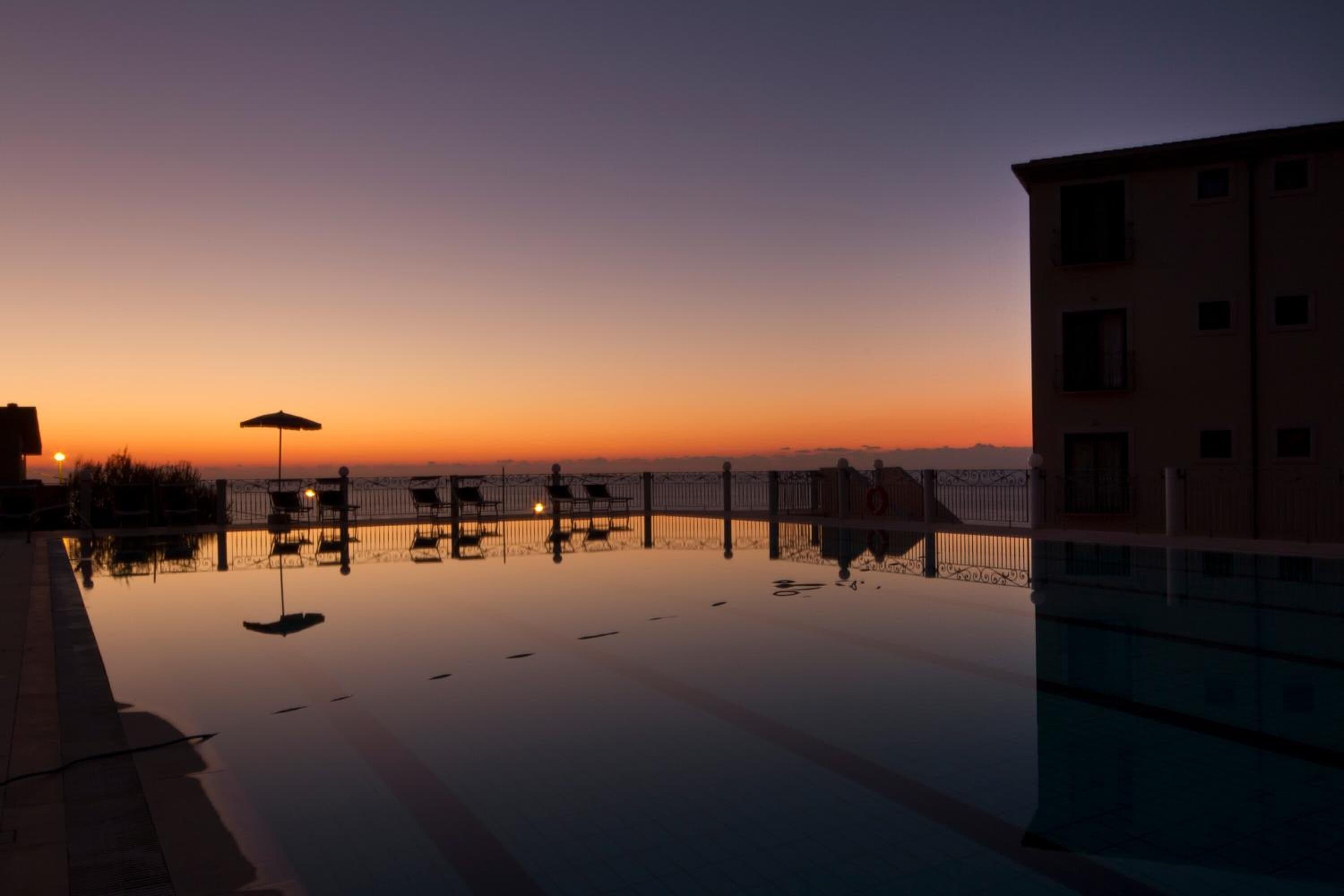 Une piscine paisible au crépuscule, avec une vue sur le coucher de soleil, une clôture et un bâtiment en arrière-plan.