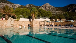 A swimming pool with clear blue water, surrounded by lounge chairs and umbrellas, with mountains in the background. - Hotel Brancamaria (Dorgali)