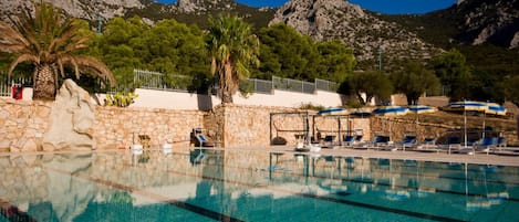 A swimming pool with clear blue water, surrounded by lounge chairs and umbrellas, with mountains in the background.