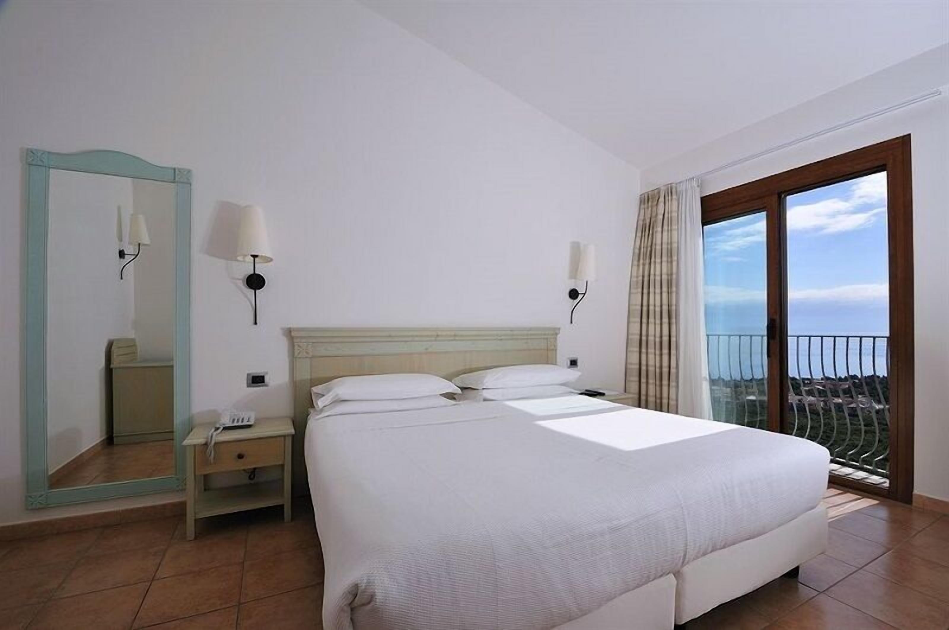 A hotel room with a large bed, a nightstand, a mirror, two wall-mounted lamps, and a balcony with a view.