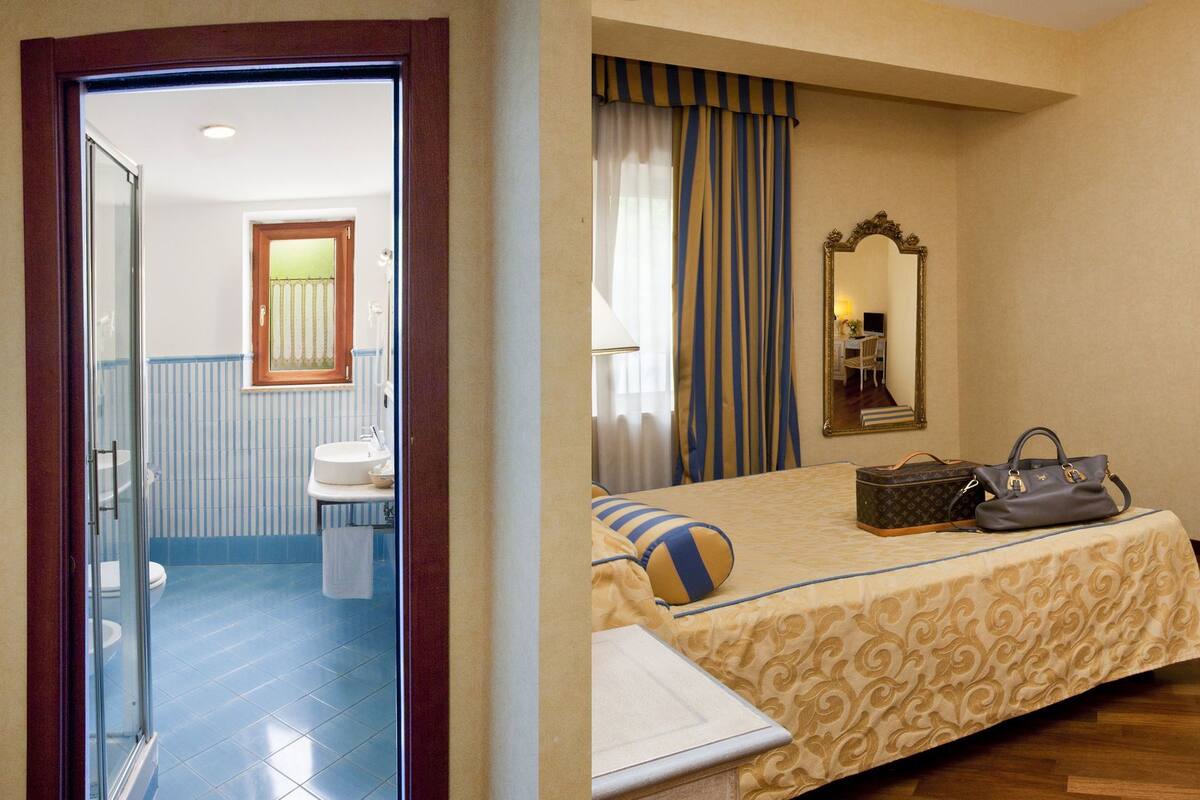 Camera d'albergo con un letto, una scrivania, una sedia, uno specchio e un bagno visibile attraverso una porta aperta.