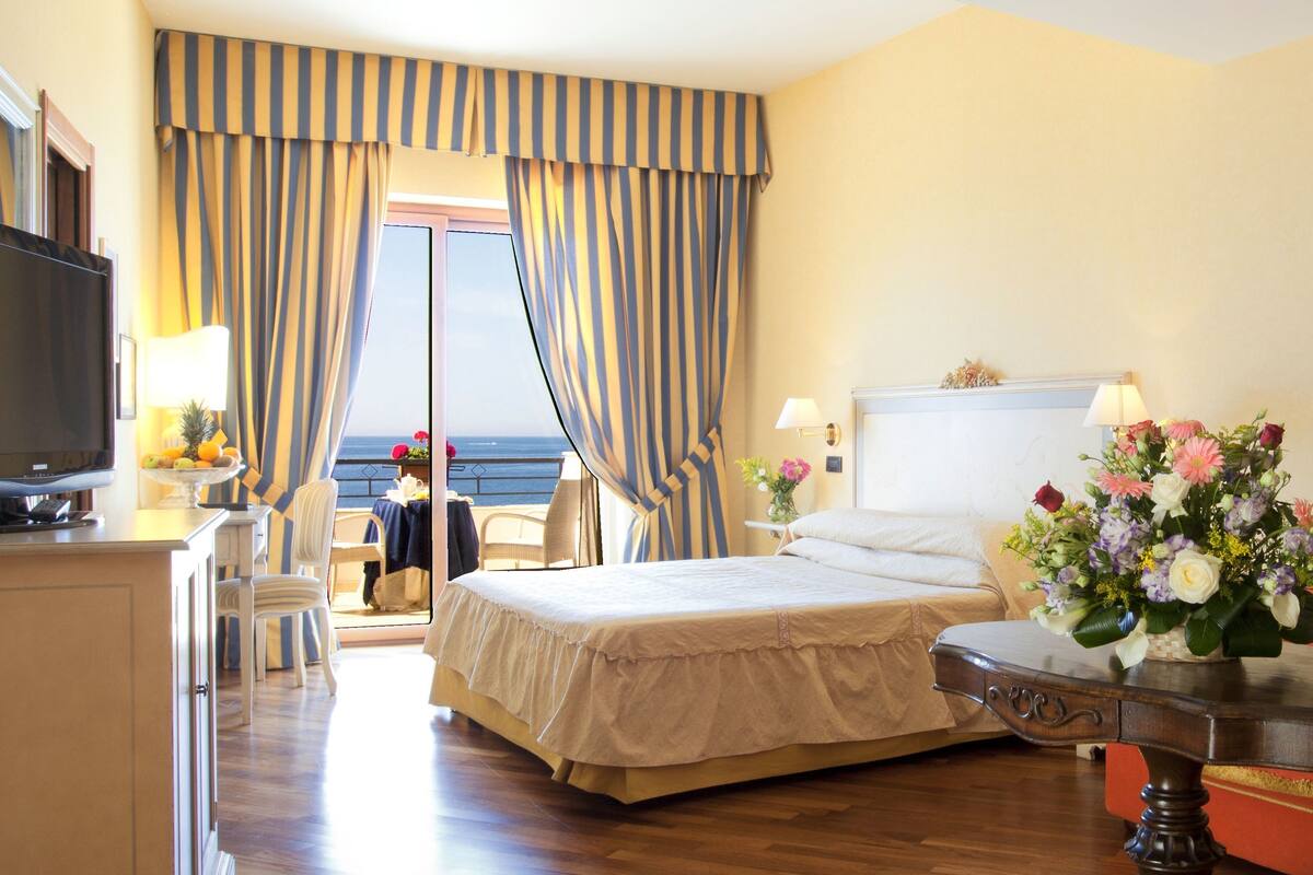 Camera d'albergo con un letto, una televisione, una scrivania, una sedia, un tavolo con un vaso di fiori e vista sul mare tra le tende.