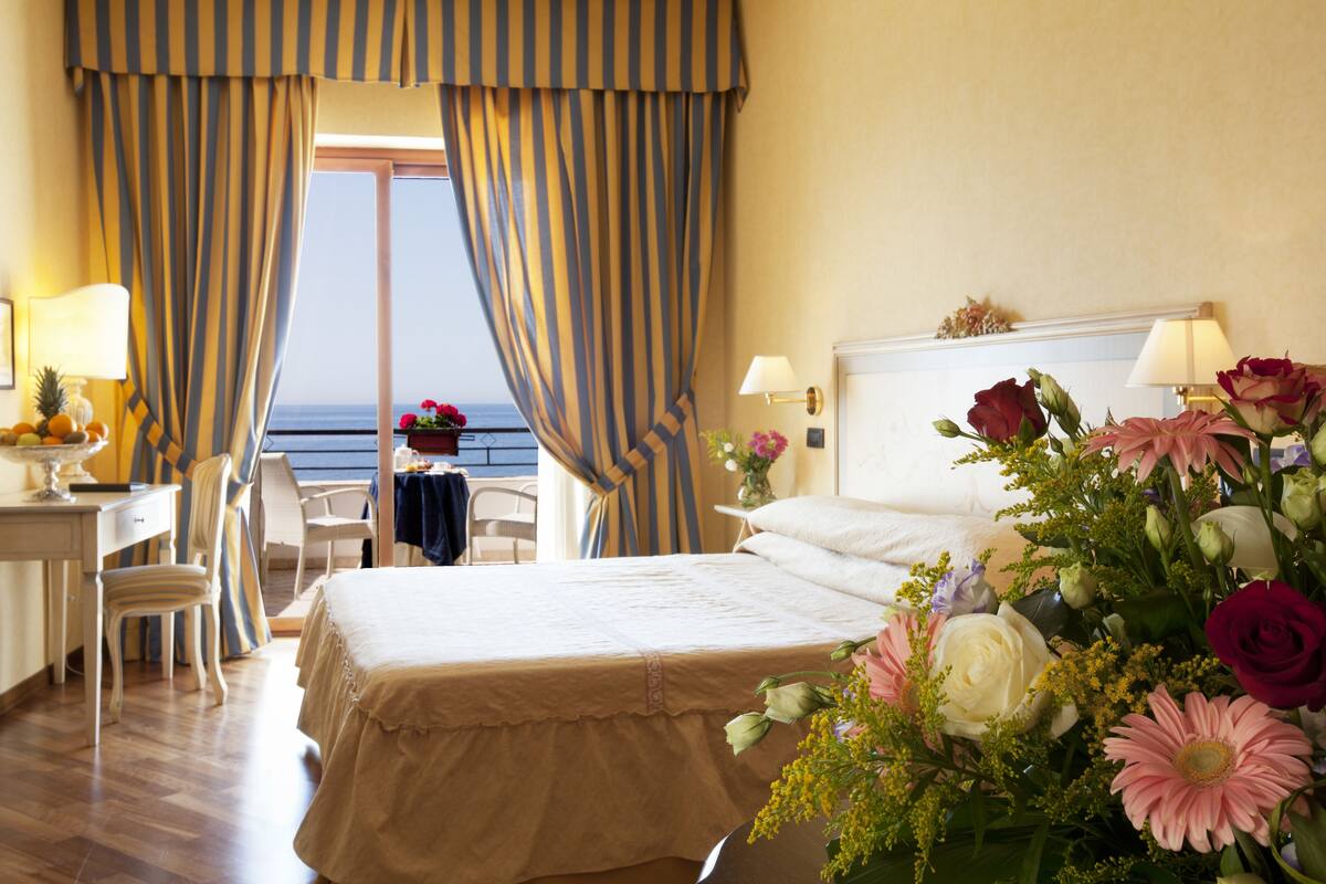 Una camera d'albergo con un letto, una scrivania, una sedia, vista sul mare e un composizione floreale.