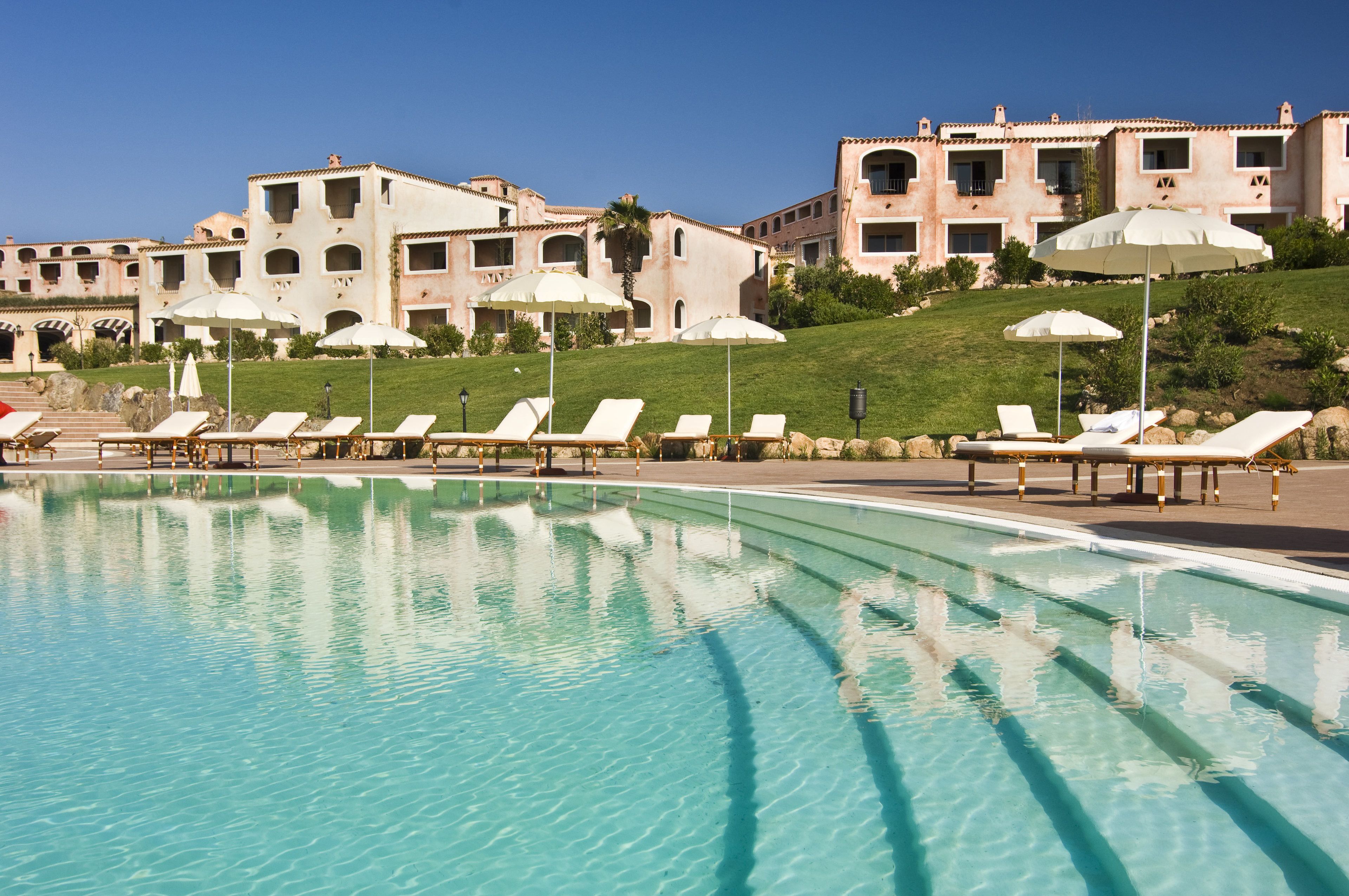 A pool with lounge chairs and umbrellas, a building with multiple balconies, and a clear sky.