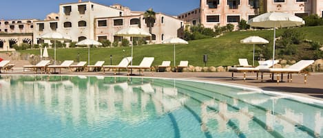 A pool with lounge chairs and umbrellas, a building with multiple balconies, and a clear sky.