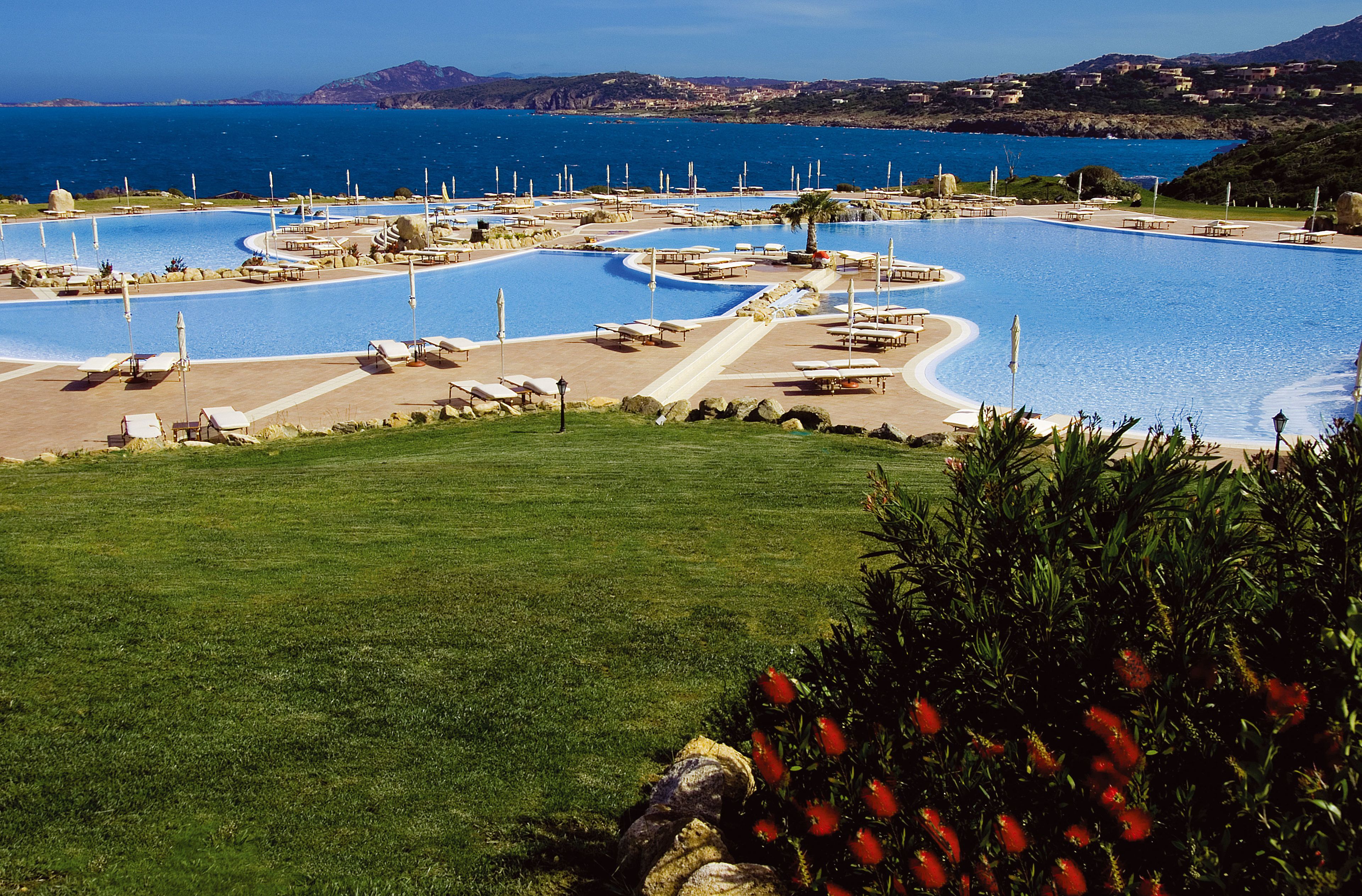 A large, winding pool with multiple sun loungers and a palm tree, surrounded by a well-maintained lawn and a view of the sea.