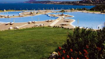 A large, winding pool with multiple sun loungers and a palm tree, surrounded by a well-maintained lawn and a view of the sea.