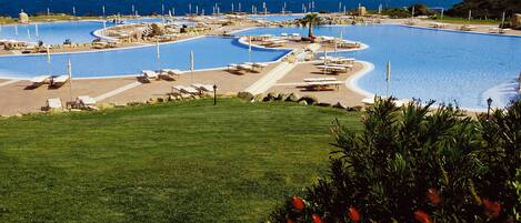 A large, winding pool with multiple sun loungers and a palm tree, surrounded by a well-maintained lawn and a view of the sea.