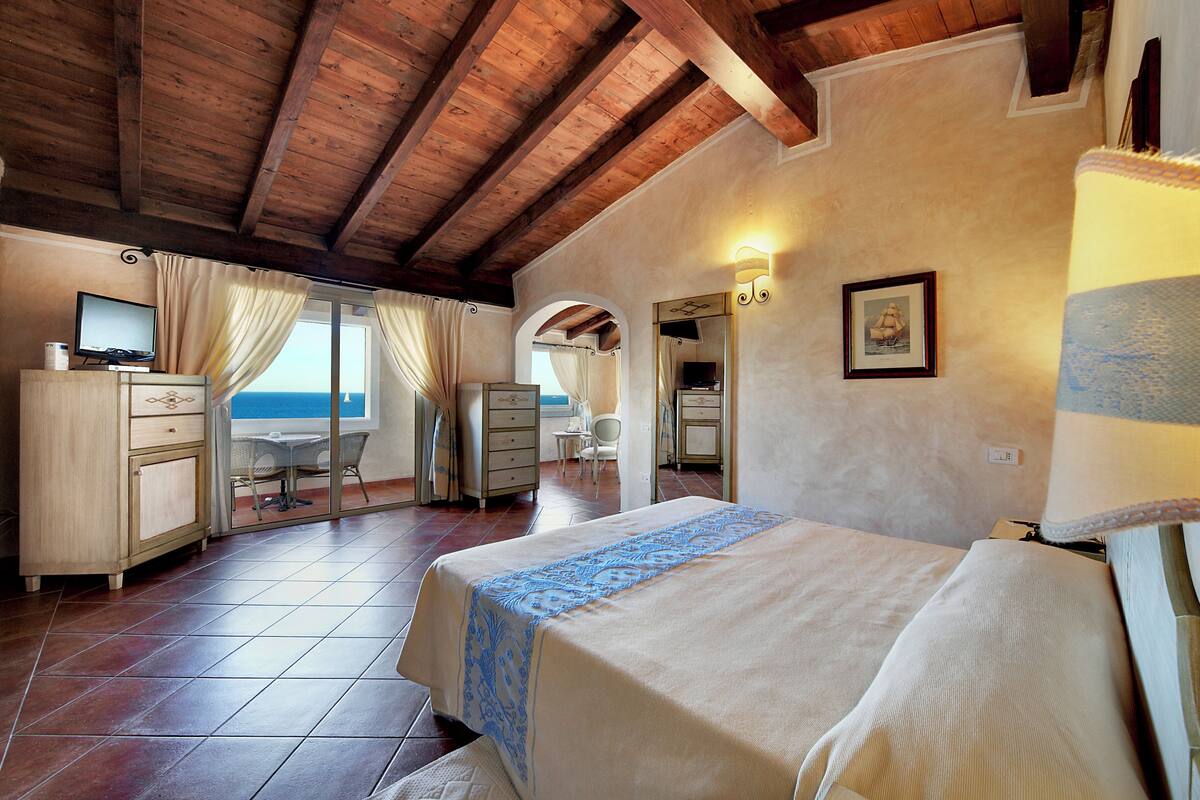 Une chambre avec un lit, un bureau et une chaise, une salle de bain avec un miroir et un lavabo, et une vue sur la mer par une fenêtre.