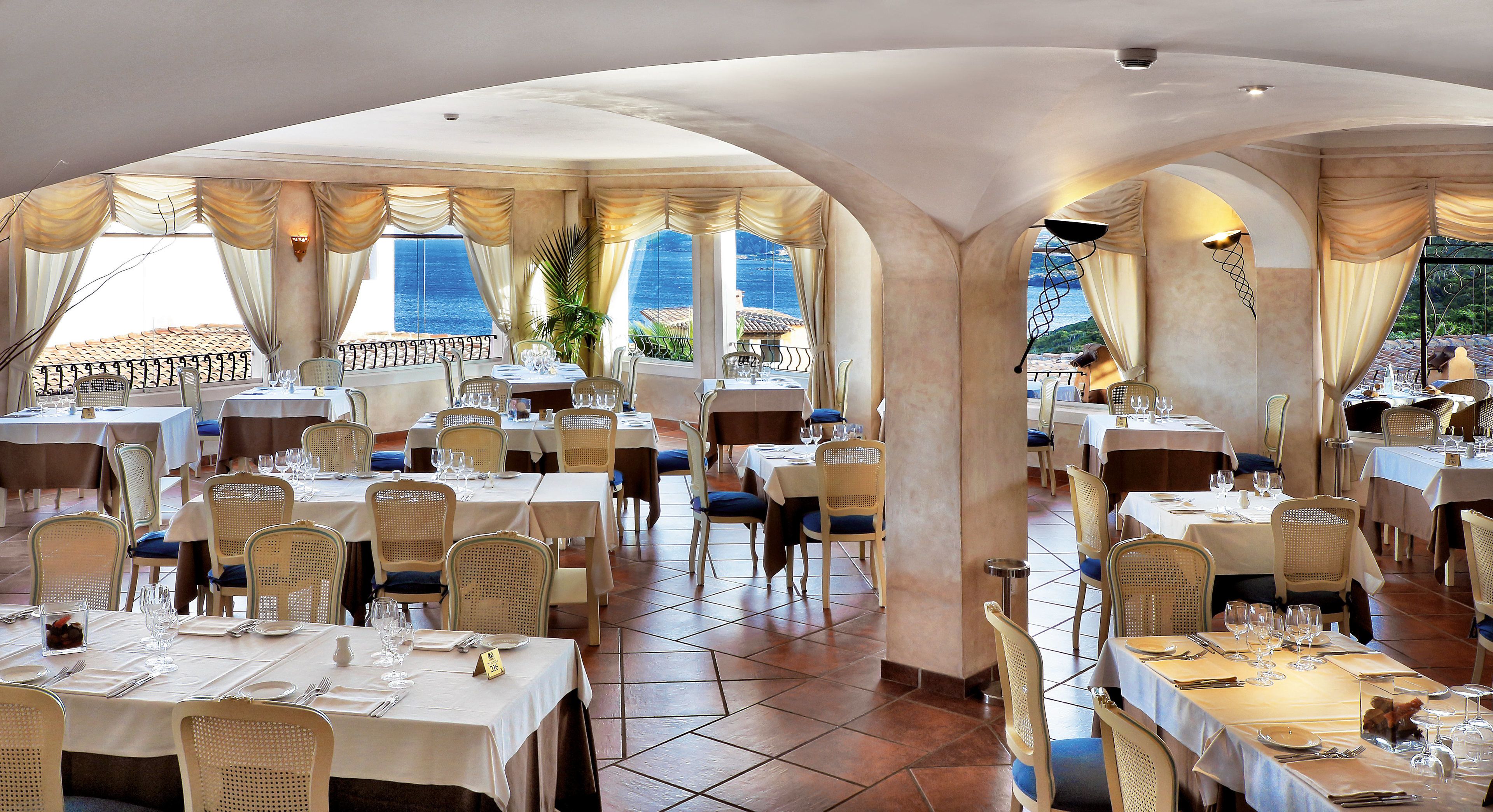 A restaurant with multiple tables and chairs, arranged in a spacious interior with large windows offering a view of the sea.