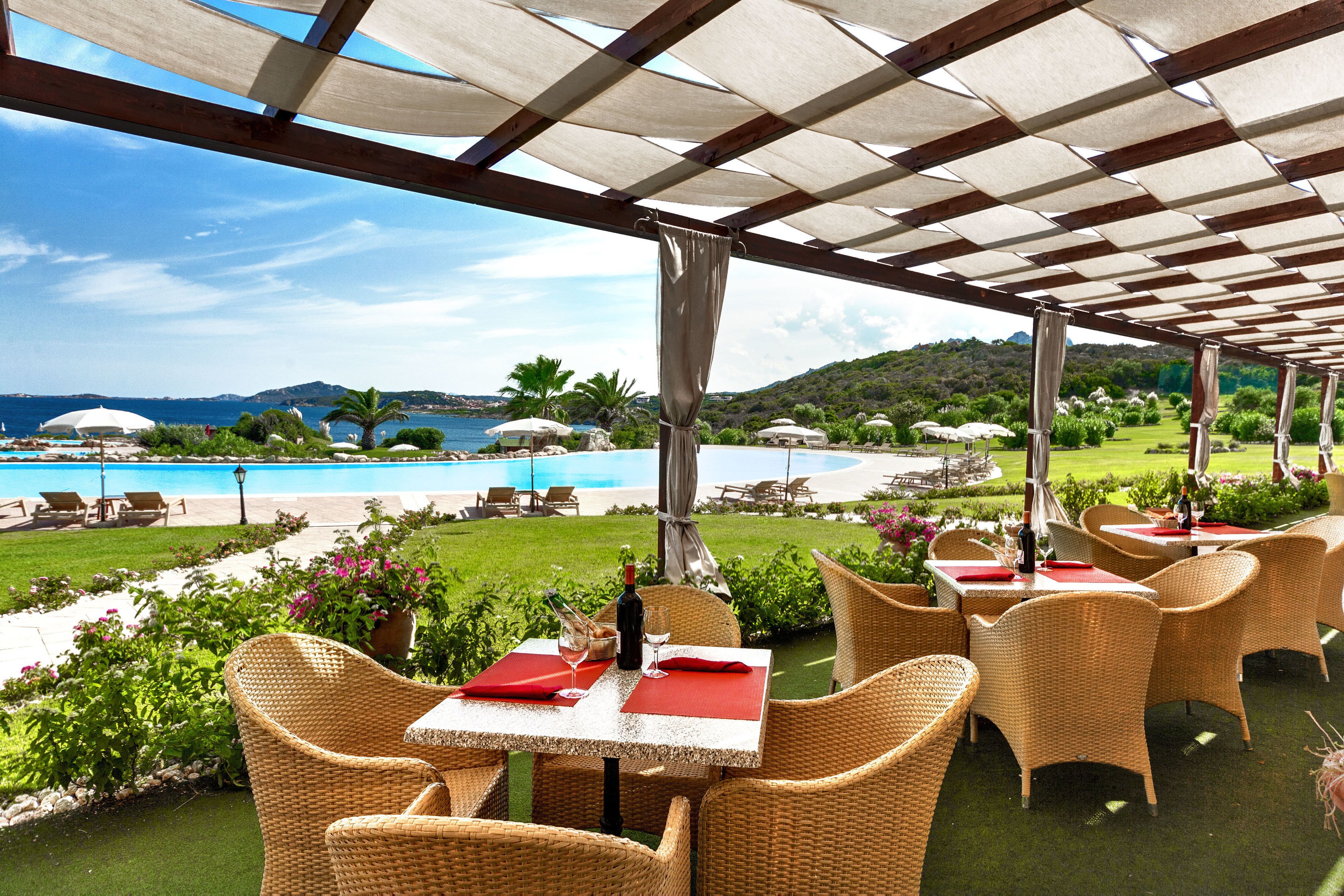 An outdoor patio with wicker furniture, tables with red napkins, and a view of a pool and greenery.