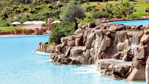 Une piscine agrémentée d’une cascade artificielle entourée de rochers, avec des chaises longues et des parasols.