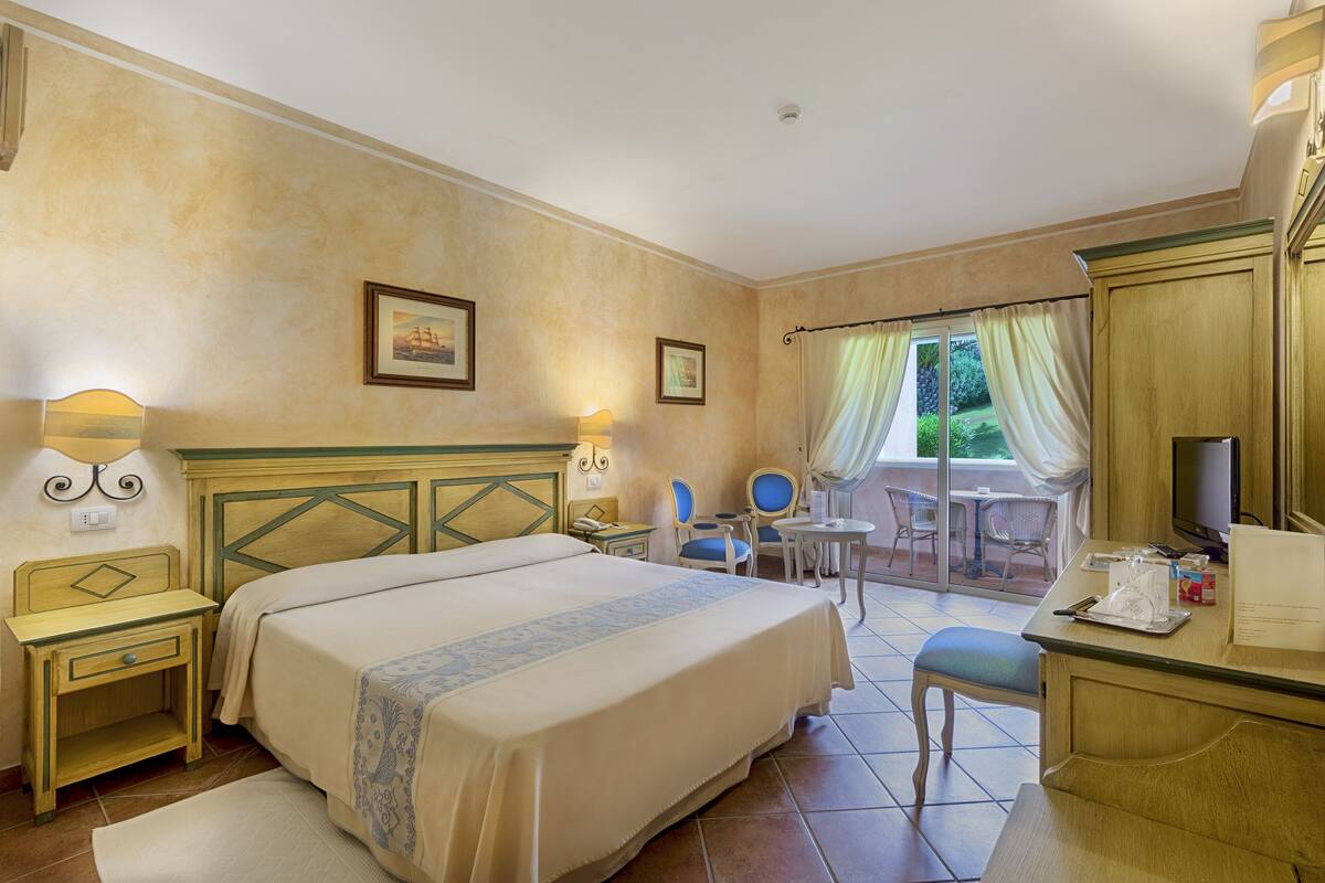 Une chambre d’hôtel avec un grand lit, un bureau, une chaise et une petite table. Il y a une fenêtre avec des rideaux et une vue sur la verdure à l’extérieur.