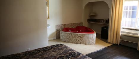 A bedroom with a large bed, a red jacuzzi, and a window with curtains.