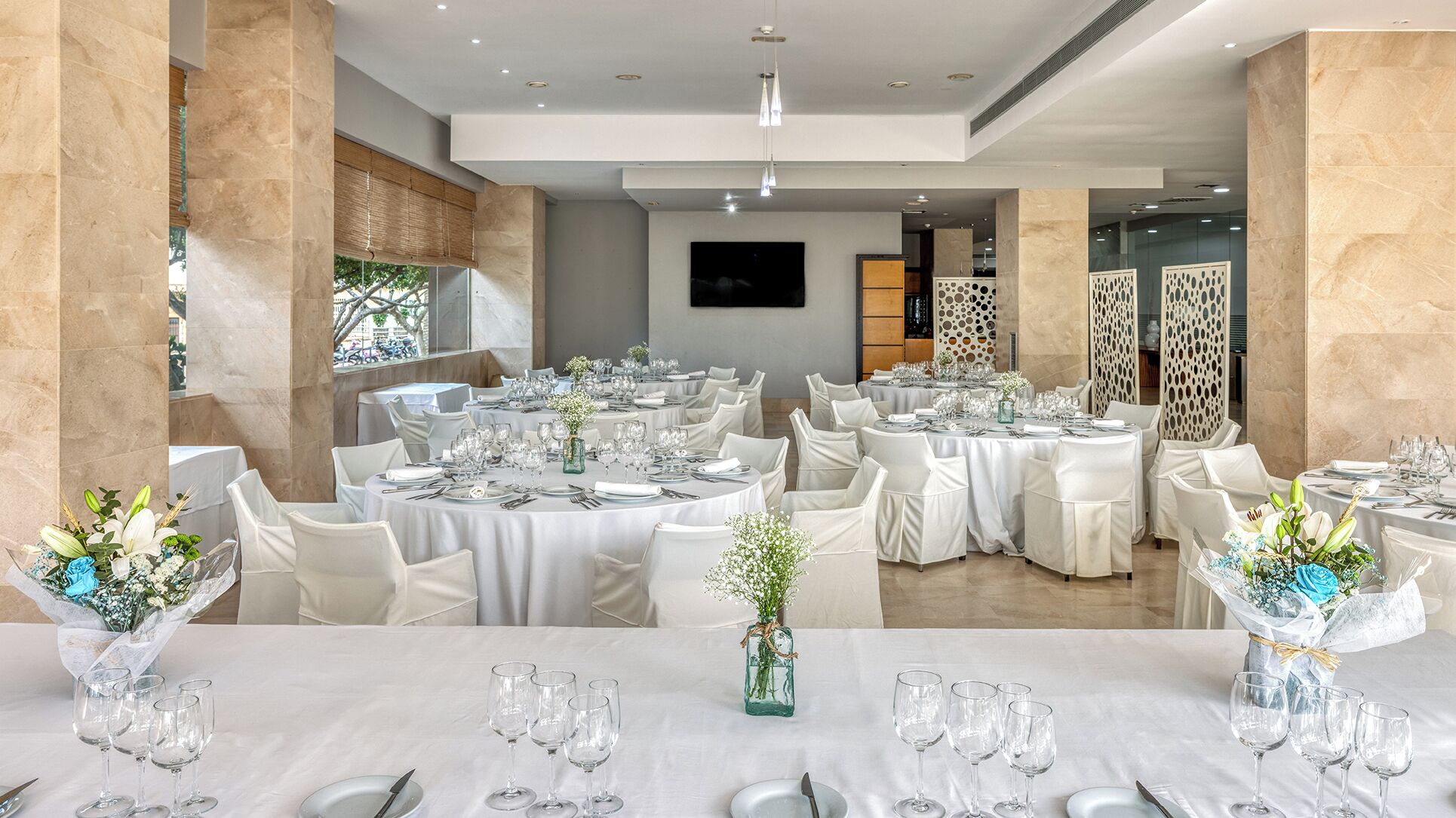 Een feestzaal met ronde tafels gedekt voor een diner, witte stoelen en bloemstukken als decoratie.