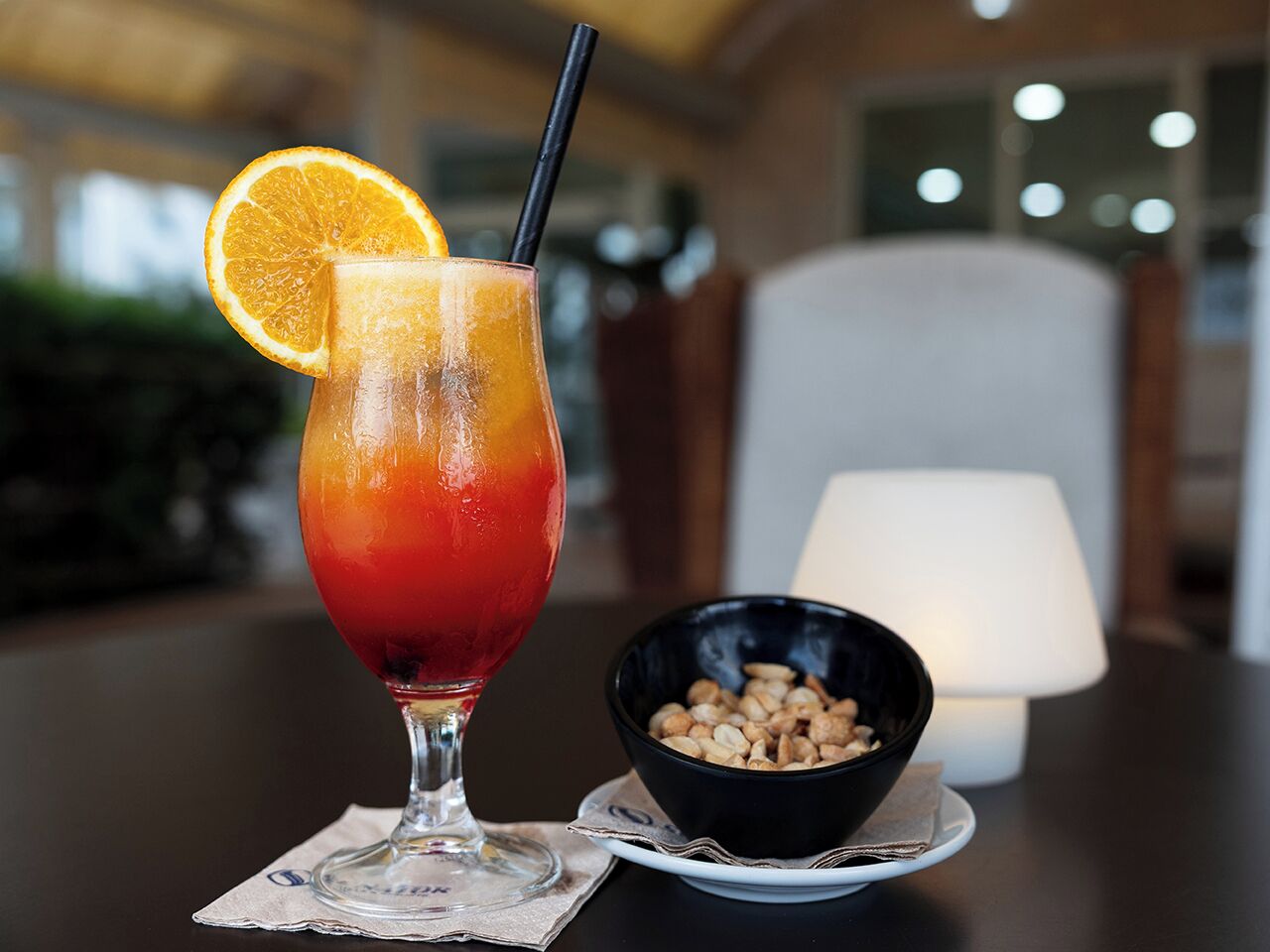 A glass of red drink with an orange slice garnish and a straw, a bowl of nuts, and a lamp in the background.
