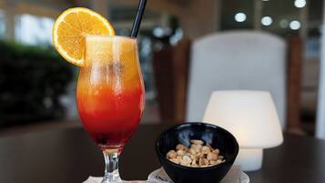 Ein Glas rotes Getränk mit Orangenscheibe und Strohhalm, eine Schüssel mit Nüssen und eine Lampe im Hintergrund.