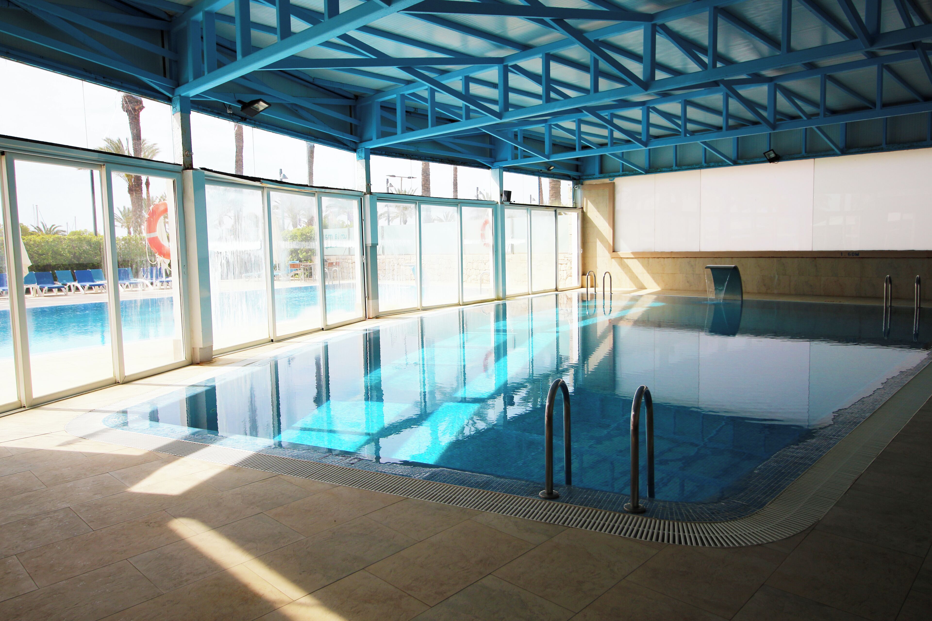 Une piscine intérieure avec des parois en verre transparent, un sol carrelé et un plafond en métal bleu.