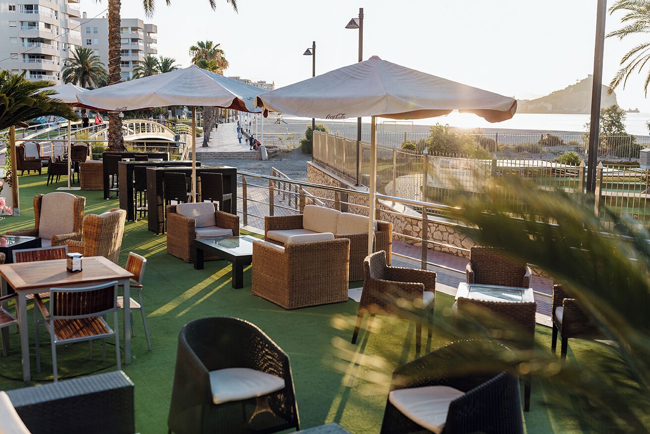 An outdoor seating area with wicker furniture and umbrellas, overlooking a coastal area with a boat and buildings in the background.