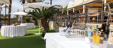 An outdoor dining area with round tables covered in white tablecloths, umbrellas, and a variety of bottles and glasses.