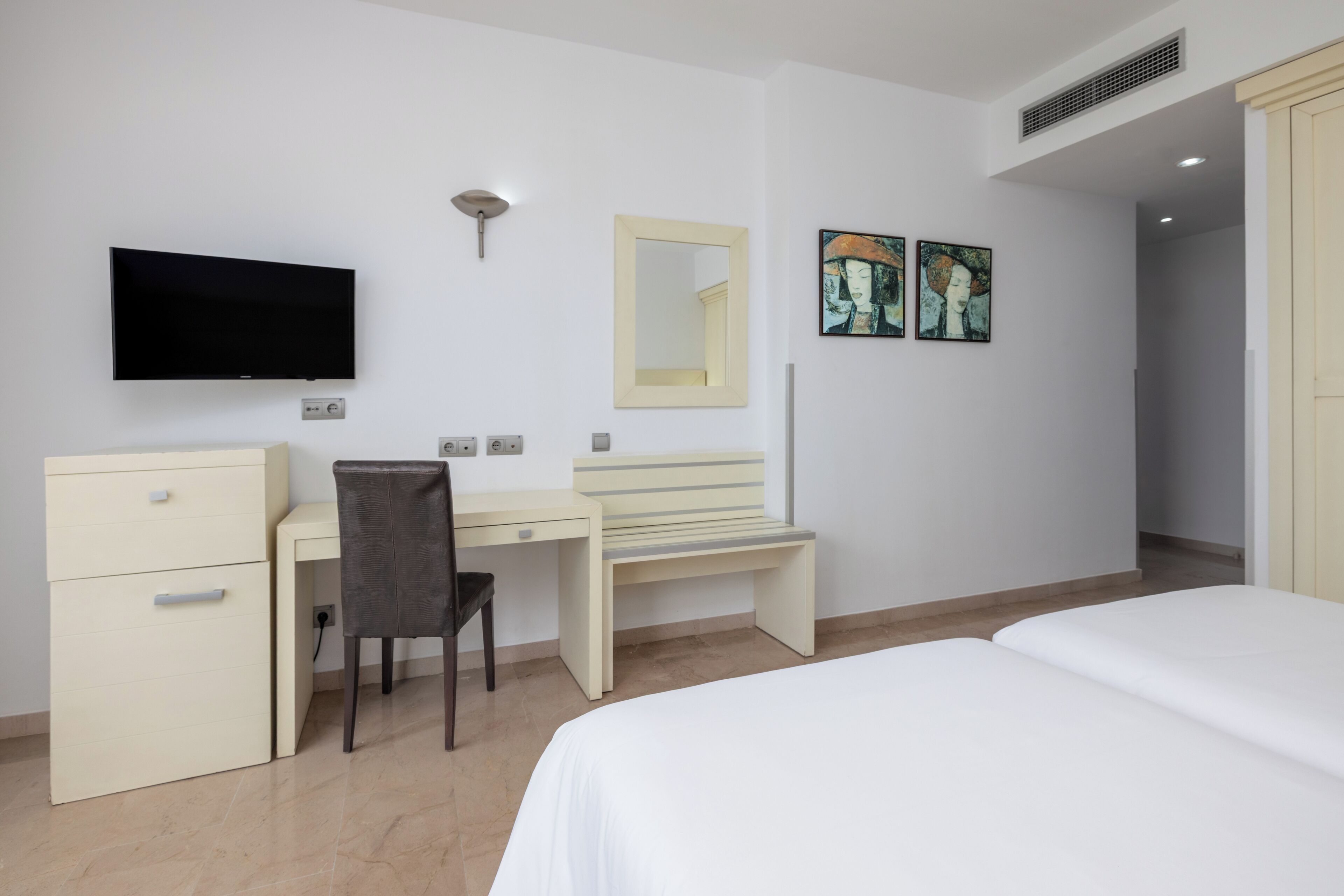 Una habitación de hotel con cama, escritorio con silla, televisión, espejo y obras de arte en la pared.