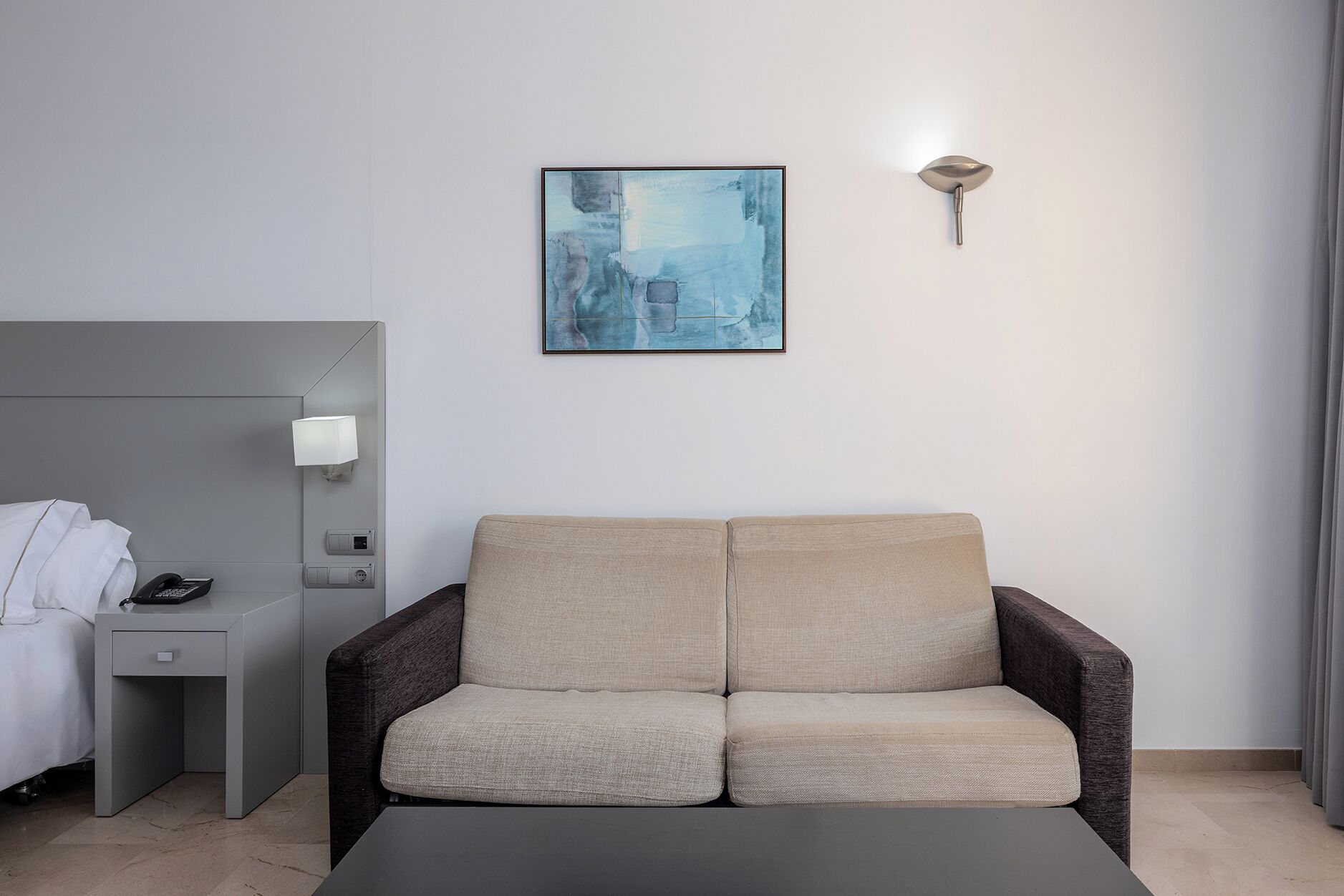 Una habitación de hotel moderna con un cabecero gris, una mesita de noche con lámpara, un sofá beige y un cuadro abstracto enmarcado en la pared.