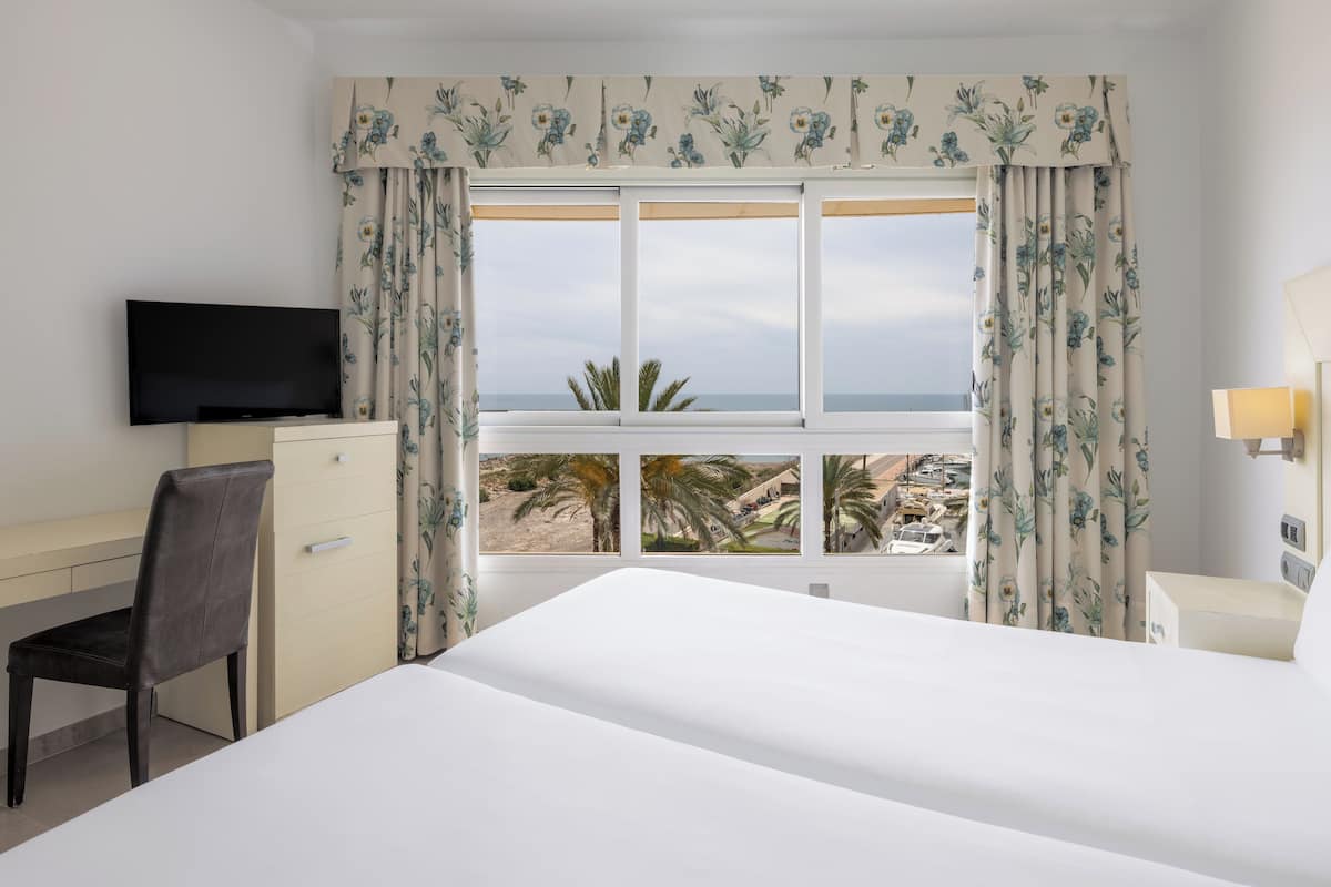 A hotel room with a bed, a desk with a TV, and a view of the beach and palm trees.