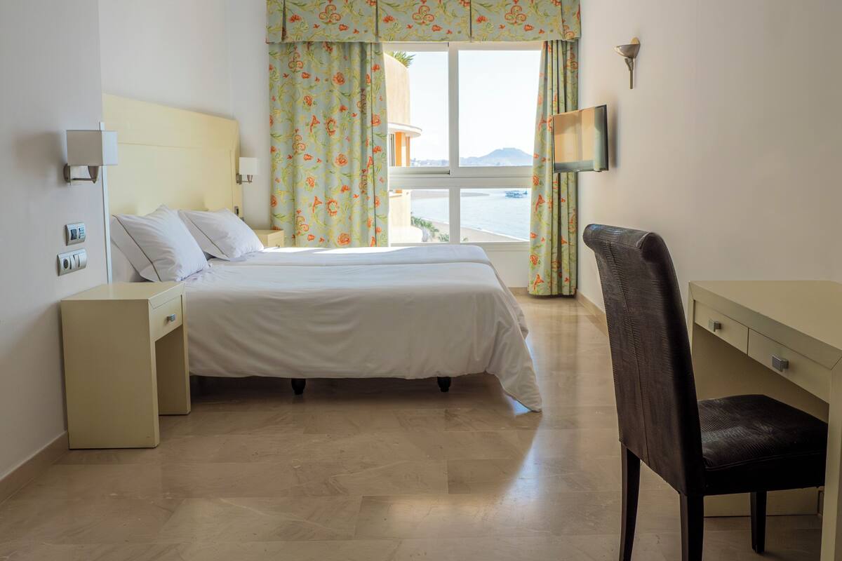 A hotel room with a bed, a desk, a chair, a TV, and a window with curtains.