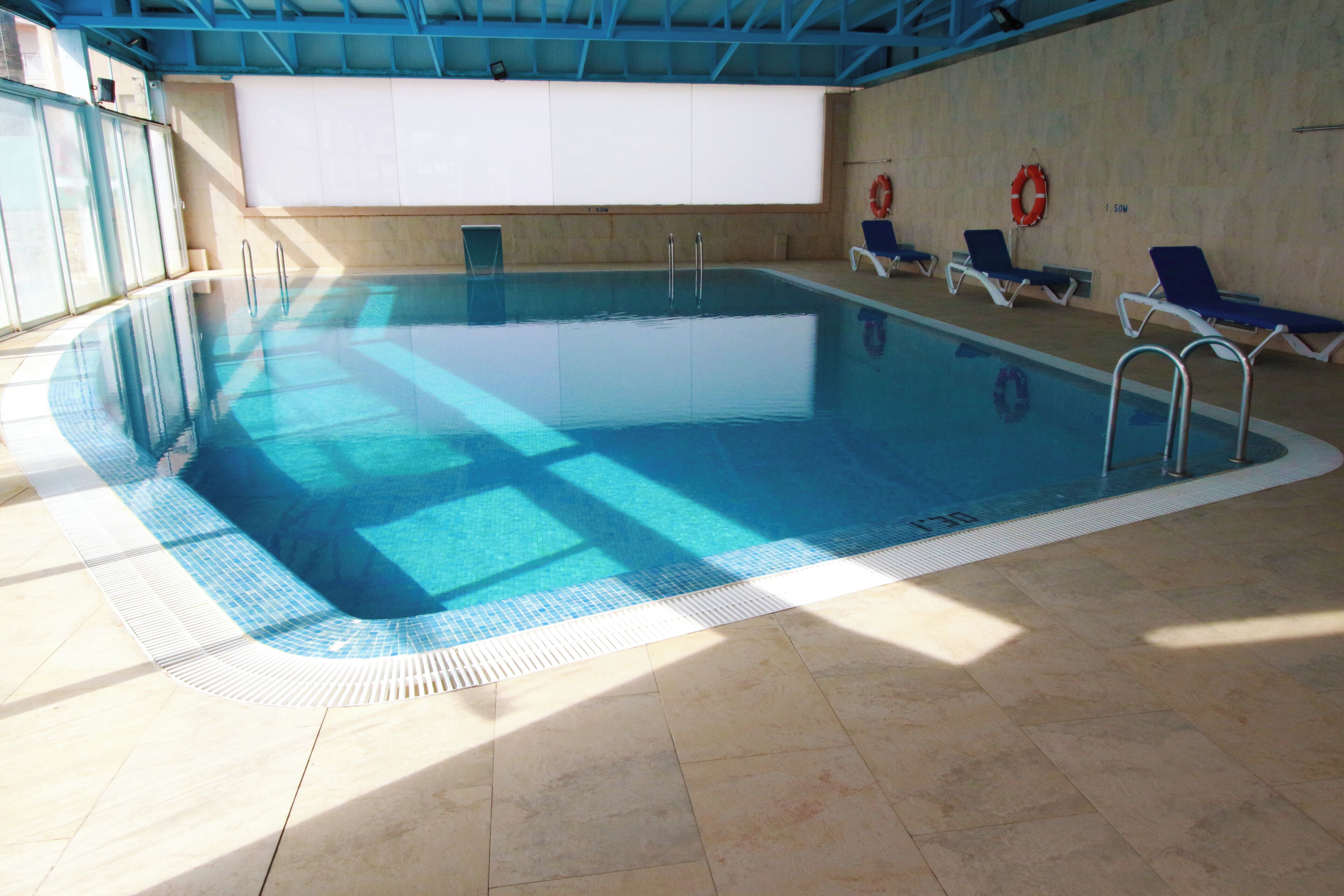 An indoor swimming pool with clear blue water, tiled floor, and sunlit walls.