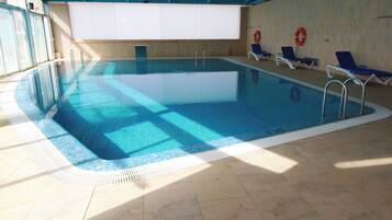 An indoor swimming pool with clear blue water, tiled floor, and sunlit walls.