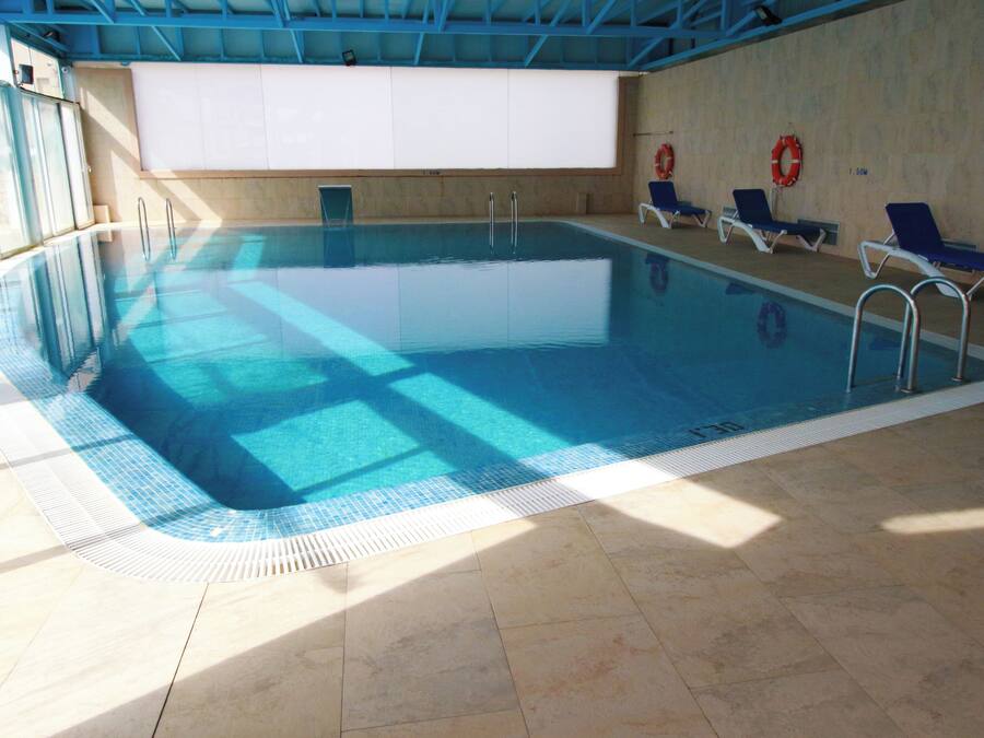 An indoor swimming pool with clear blue water, tiled floor, and sunlit walls.