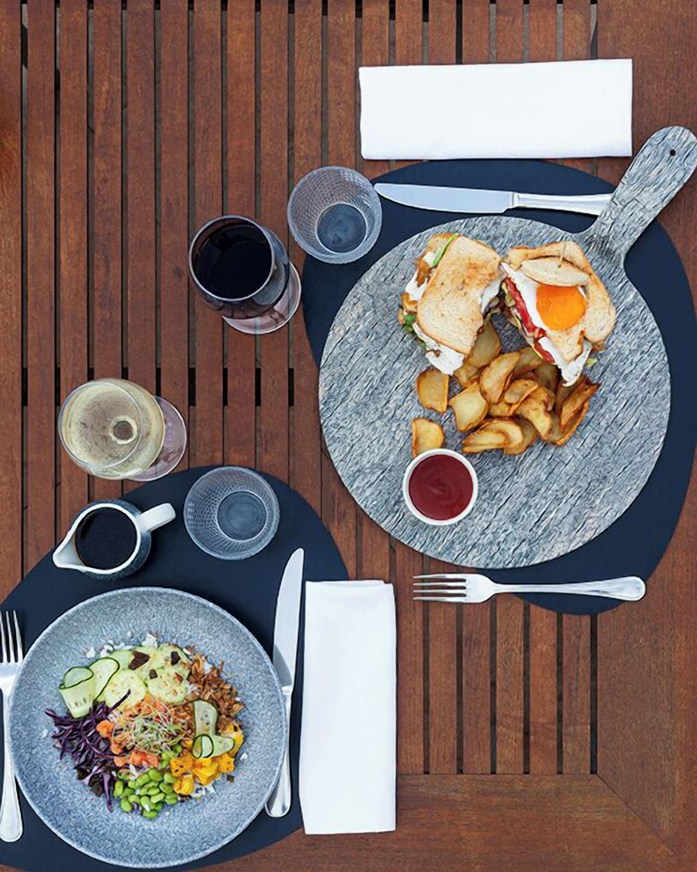 Ein Holztisch mit einer Mahlzeit aus einem Sandwich, Pommes und einem Salat, sowie zwei Gläser eines dunklen Getränks und einer kleinen Schale Soße.