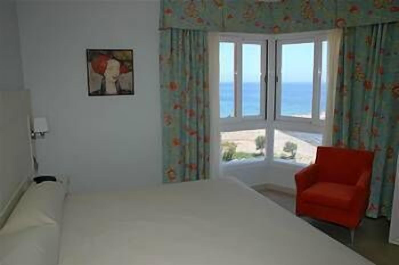 A room with a bed, a chair, a painting on the wall, and a view of the sea through a window.