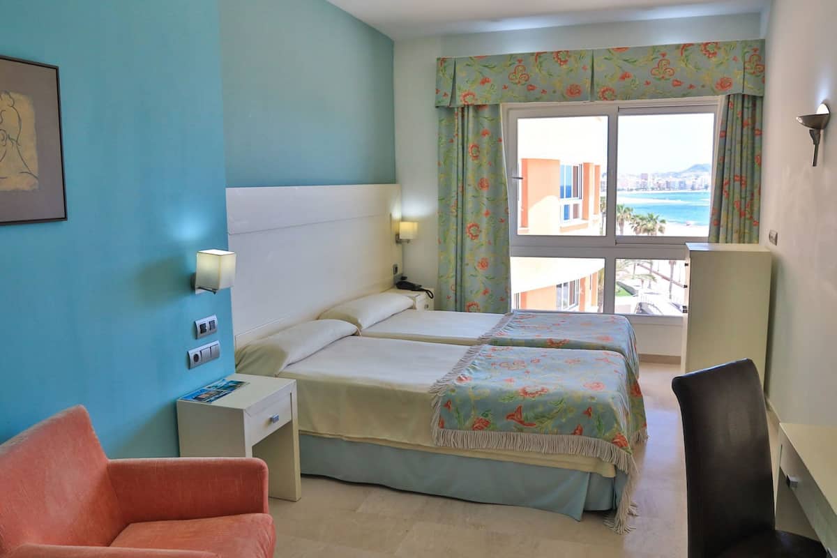 A hotel room with two beds, a desk, a chair, a window with curtains, and a view of the sea.
