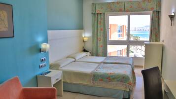 Una habitación de hotel con dos camas, un escritorio, una silla, una mesita de noche, una ventana con cortinas y una lámpara de pared.