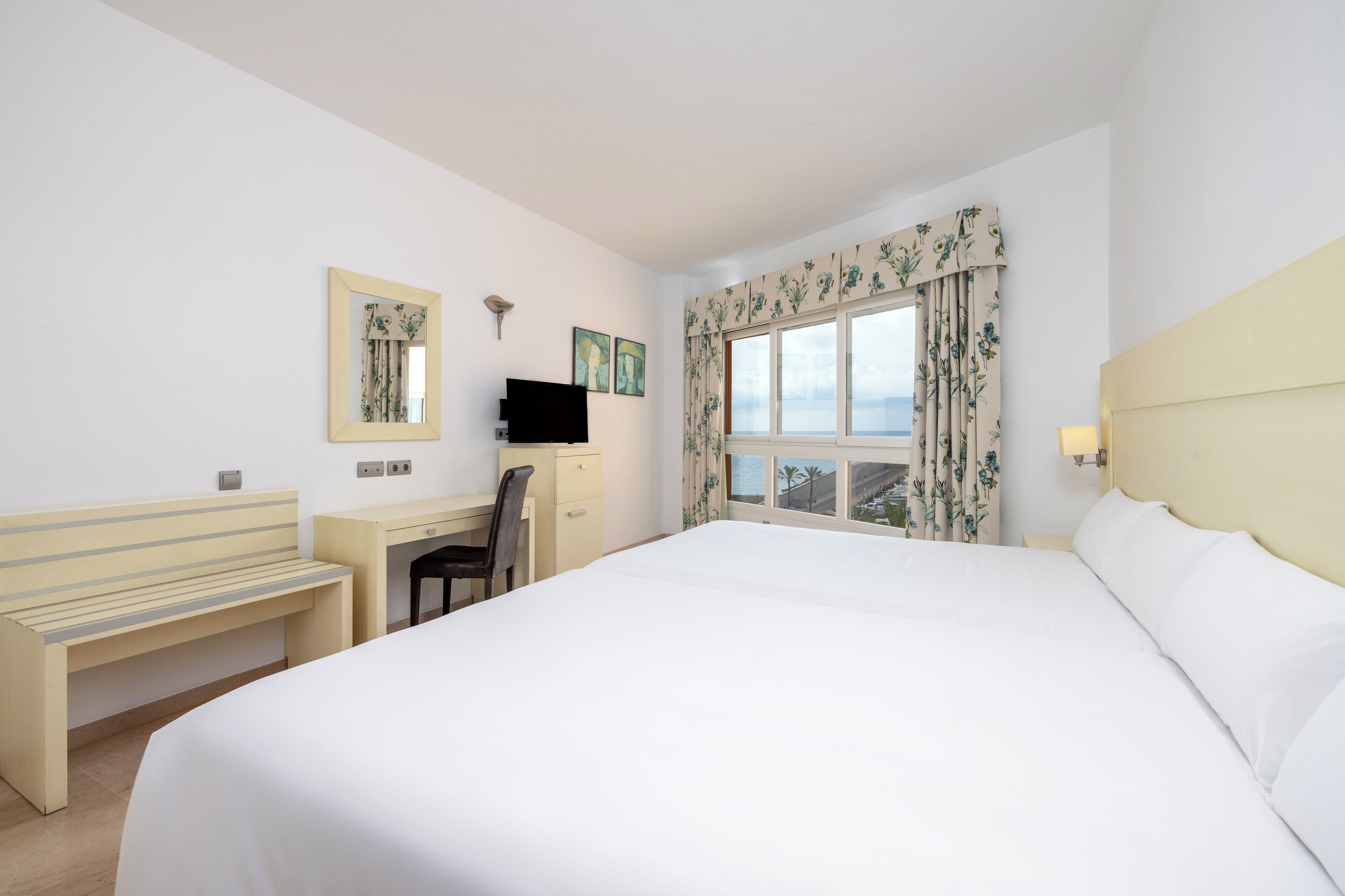 Une chambre d’hôtel avec un grand lit, un bureau avec une chaise, une petite armoire, un téléviseur à écran plat et une fenêtre agrémentée de rideaux à motifs floraux.