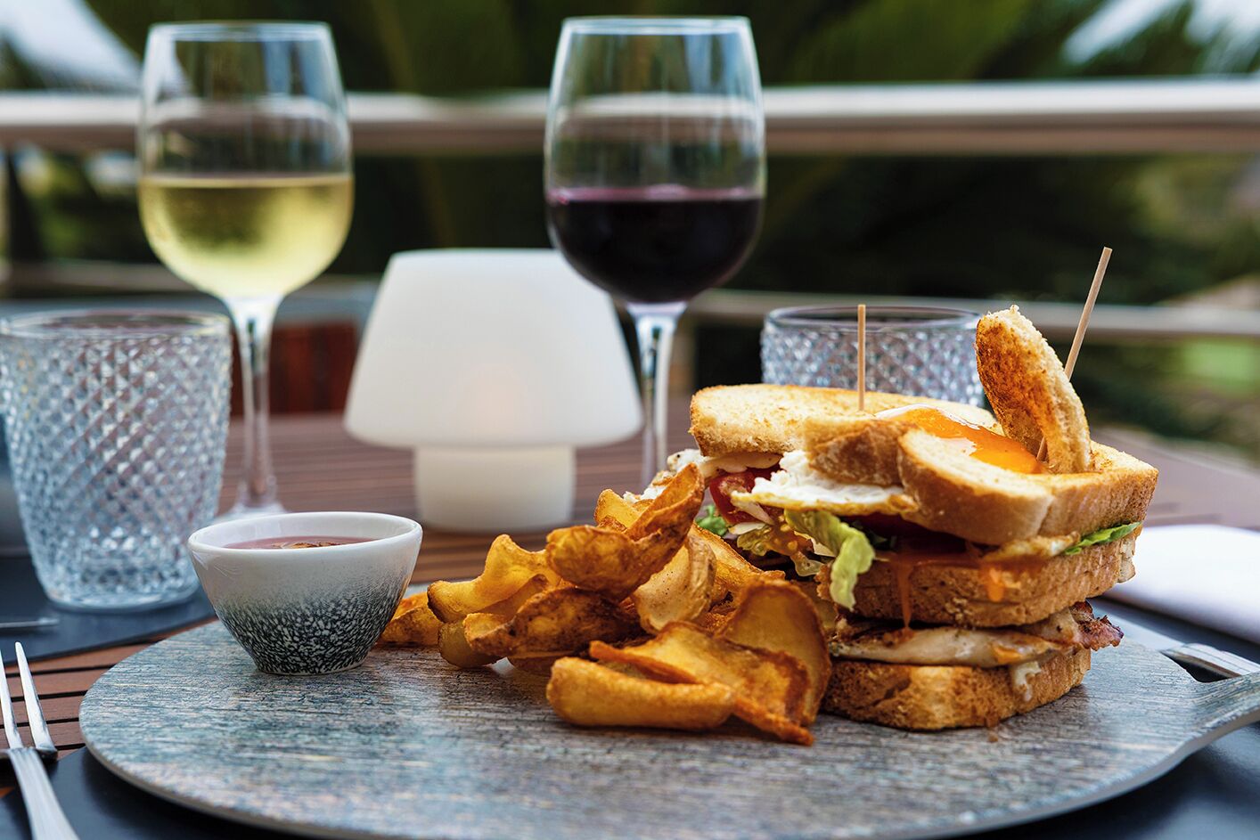 Una comida que incluye un sándwich, papas fritas y un pequeño recipiente con salsa, junto con dos copas de vino sobre una mesa.