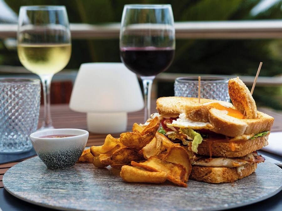 Una comida que incluye un sándwich, papas fritas y un pequeño recipiente con salsa, junto con dos copas de vino sobre una mesa.