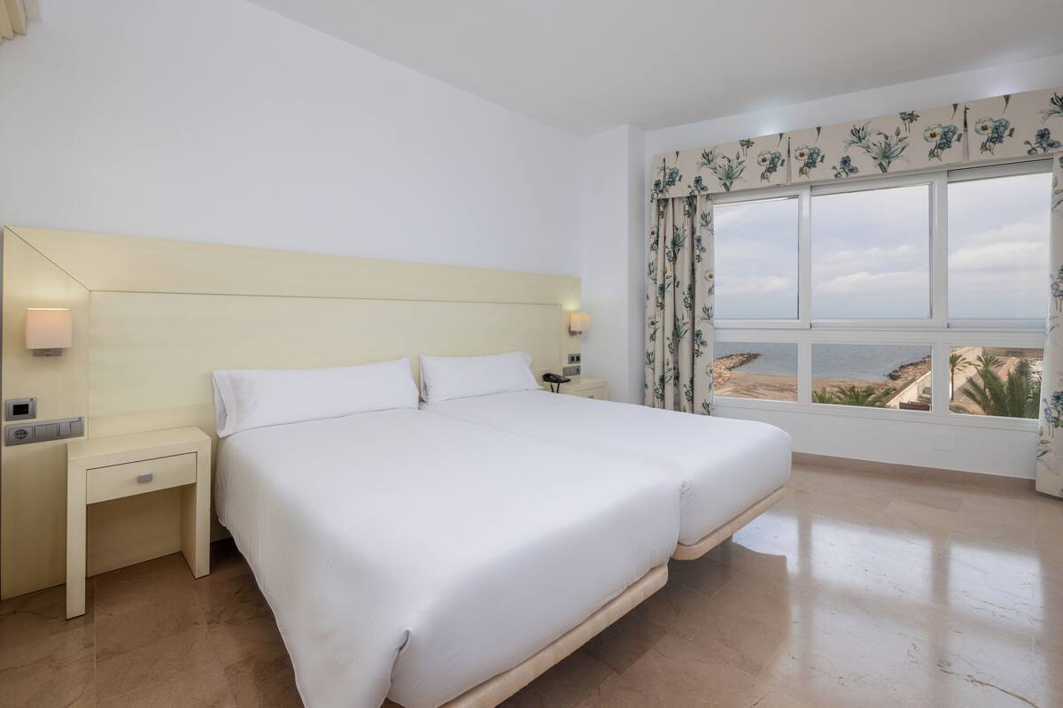 A bedroom with a large bed, a nightstand, a lamp, a window with a view of the sea, and a floral curtain.
