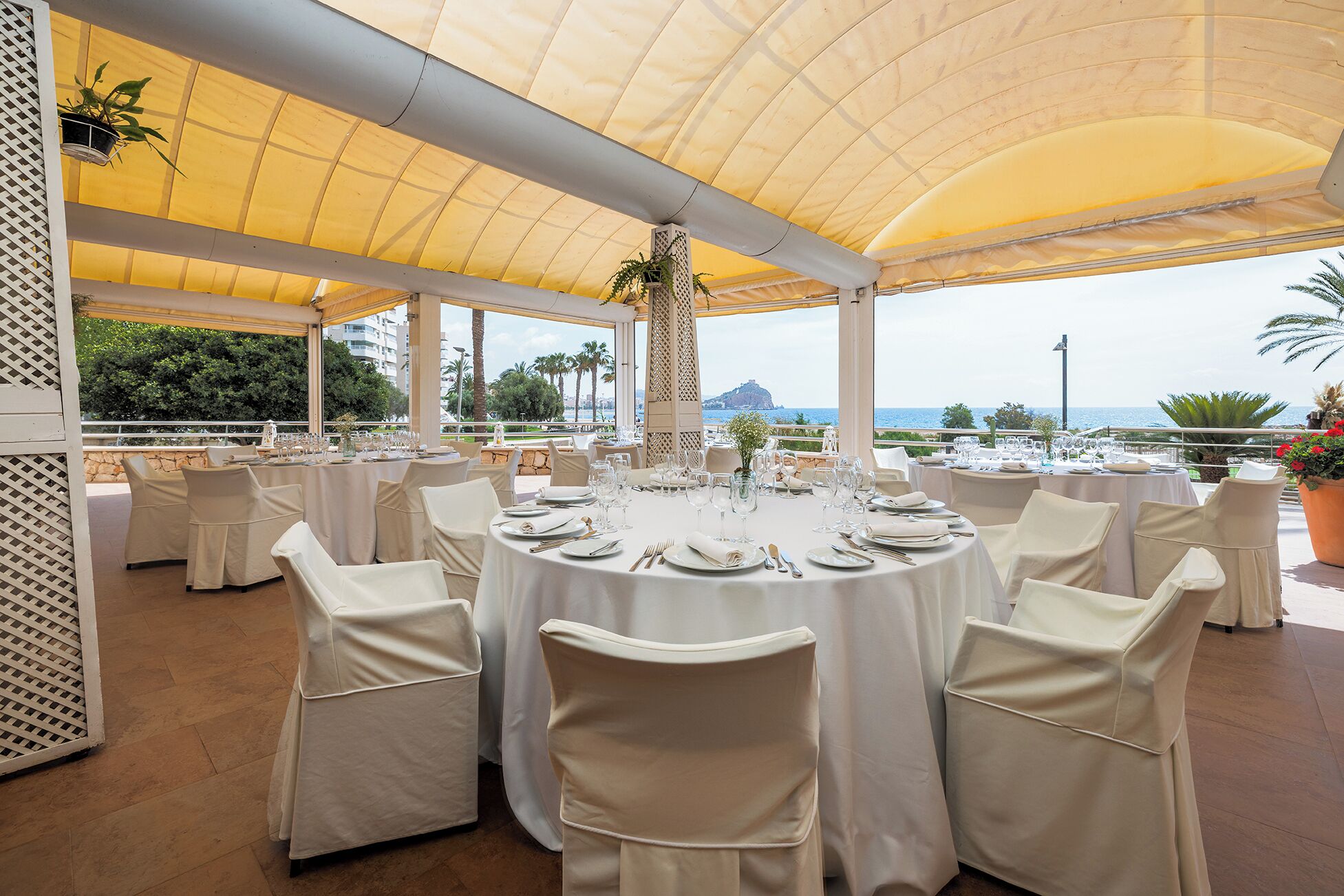 A covered outdoor dining area with round tables set for a meal, overlooking a scenic view.