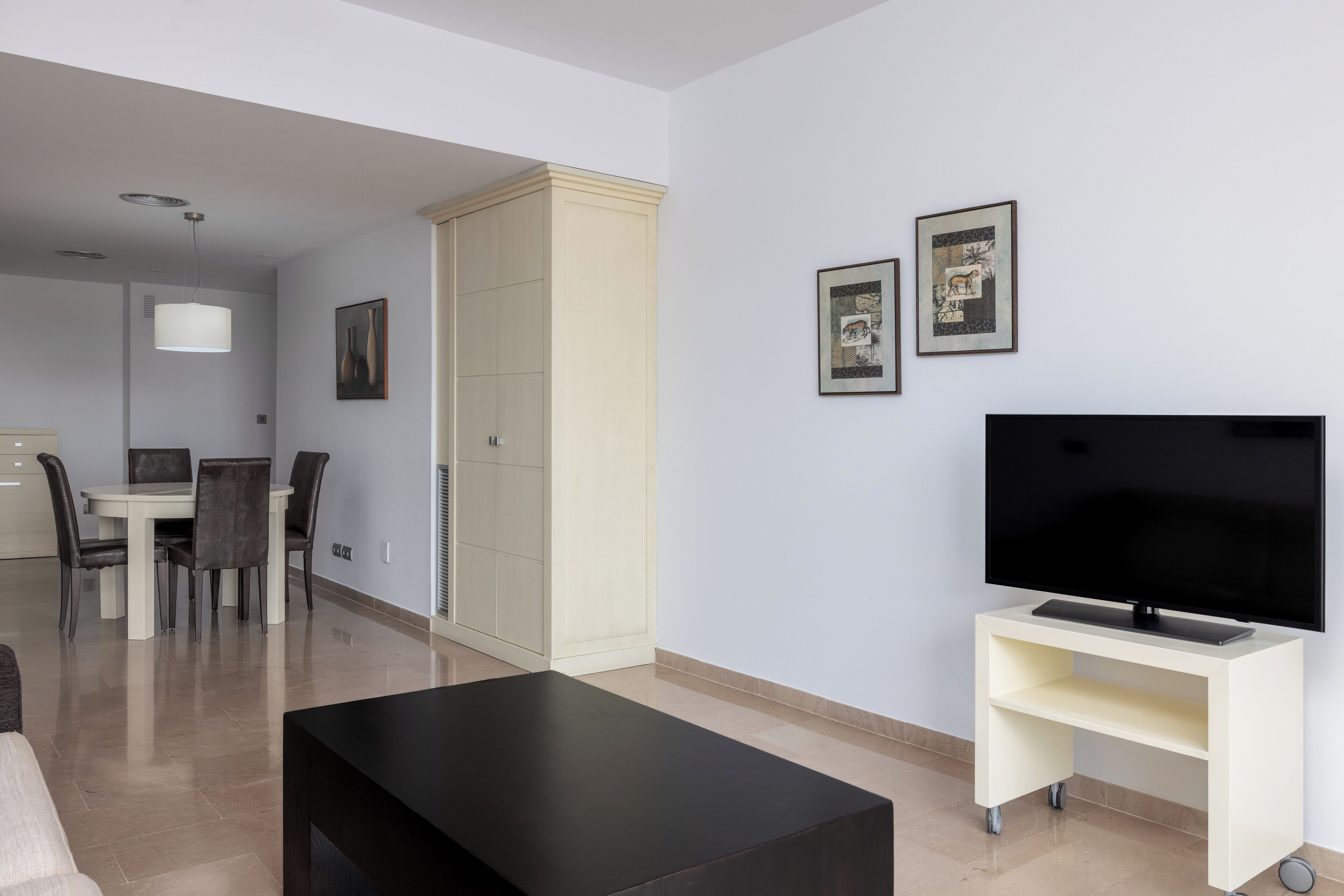 Una sala de estar moderna con una televisión de pantalla plana sobre un mueble, una zona de comedor con mesa y sillas, y un armario.