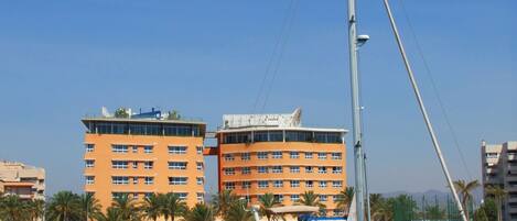 A marina with several boats docked, a tall building with multiple balconies, and palm trees in the background.