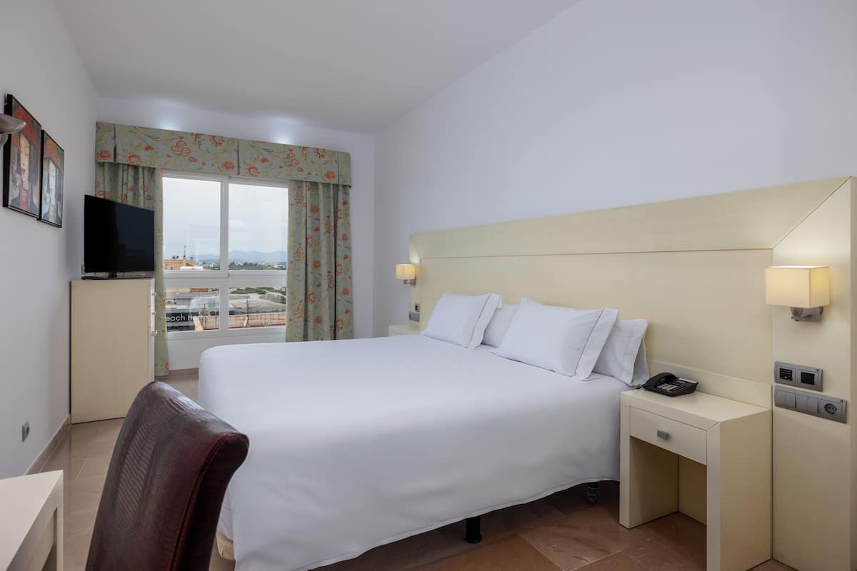 A hotel room with a large bed, a desk, a television, and a view of the city through the window.