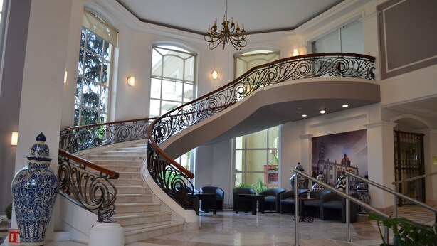 A grand staircase with ornate metal railings, a chandelier, and a seating area with modern furniture. - Plaza Camelinas Hotel (Querétaro)
