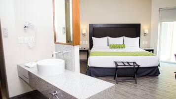 Una habitación de hotel con una cama grande, un baño con lavamanos y espejo, y un taburete pequeño.