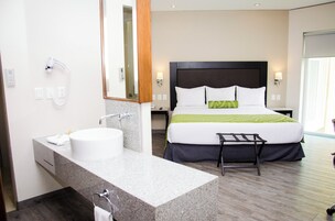 Una habitación de hotel con una cama grande, un baño con lavamanos y espejo, y un taburete pequeño.