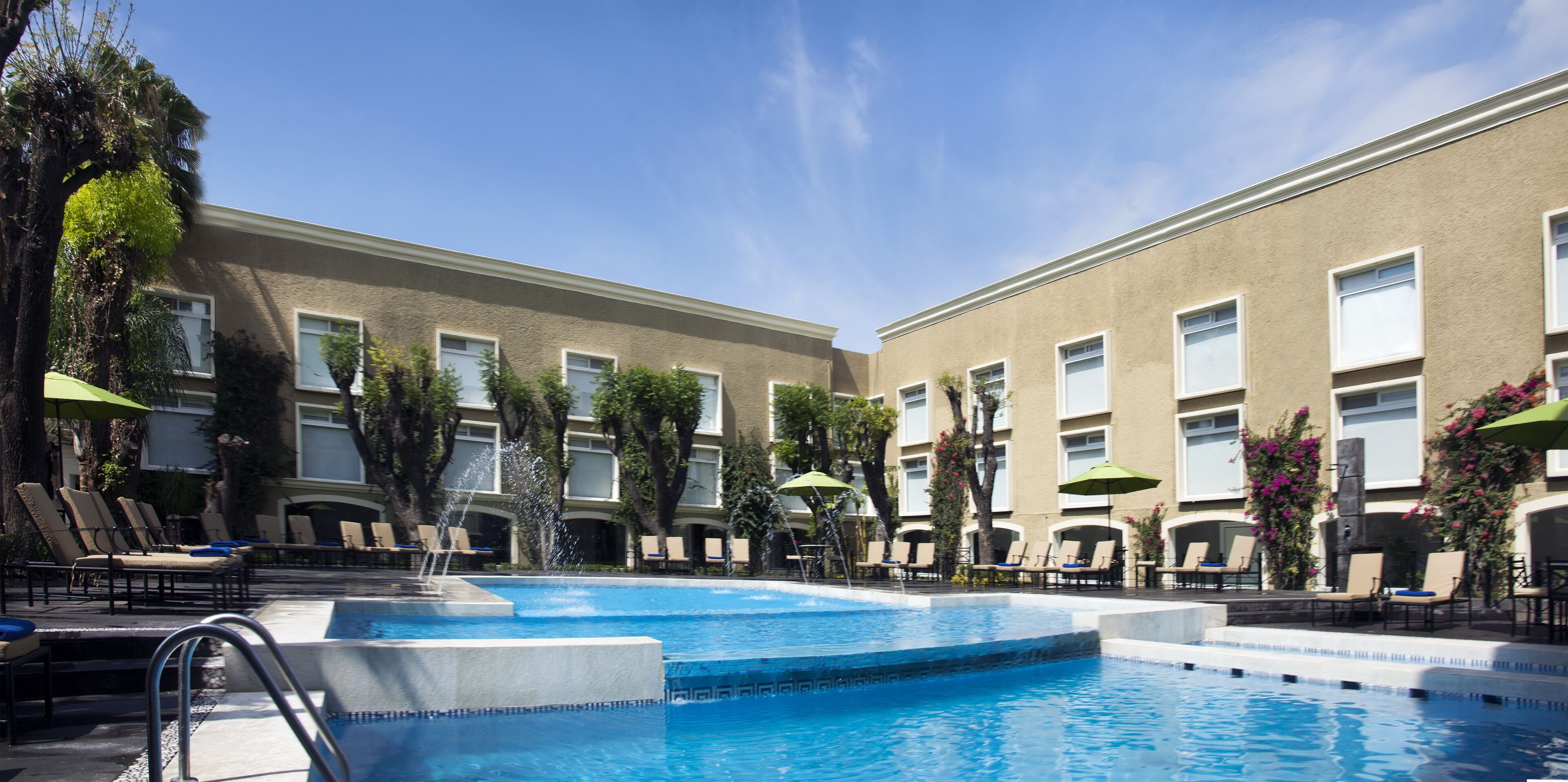 Une piscine entourée de chaises longues et de parasols, bordée d’un bâtiment aux nombreuses fenêtres, sous un ciel dégagé.