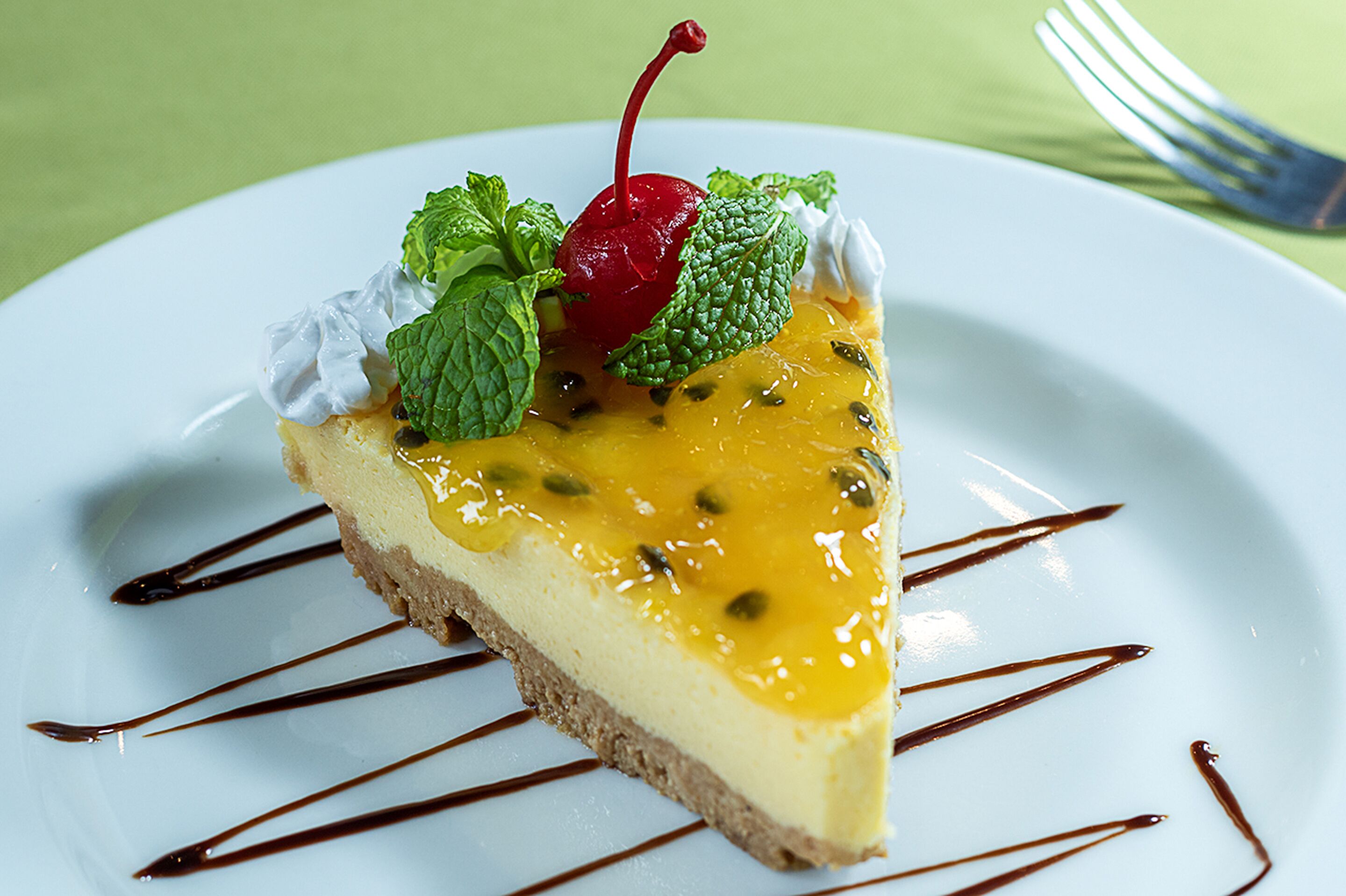 Una rebanada de pastel de queso con cereza, hojas de menta y un hilo de salsa de chocolate sobre un plato blanco.